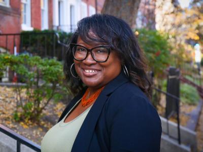 Headshot of Jameta Barlow in a green shirt with a blue jacket over it standing in front of a street of row houses.