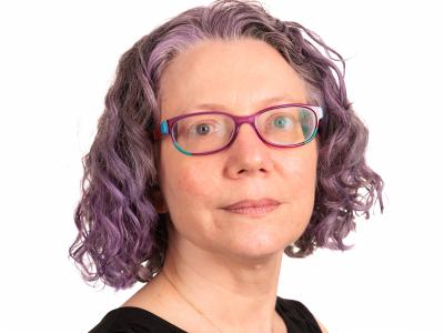 Headshot of Carol Hayes in a black shirt with a white background. Her hair is purple and very cool.
