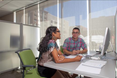 Two people sit sit by side at a desk computer talking and writing in notebooks.