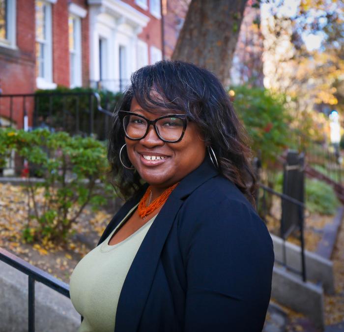 Headshot of Jameta Barlow in a green shirt with a blue jacket over it standing in front of a street of row houses.