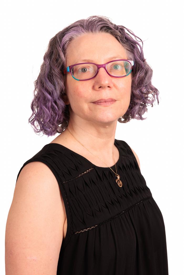 Headshot of Carol Hayes in a black shirt with a white background. Her hair is purple and very cool.