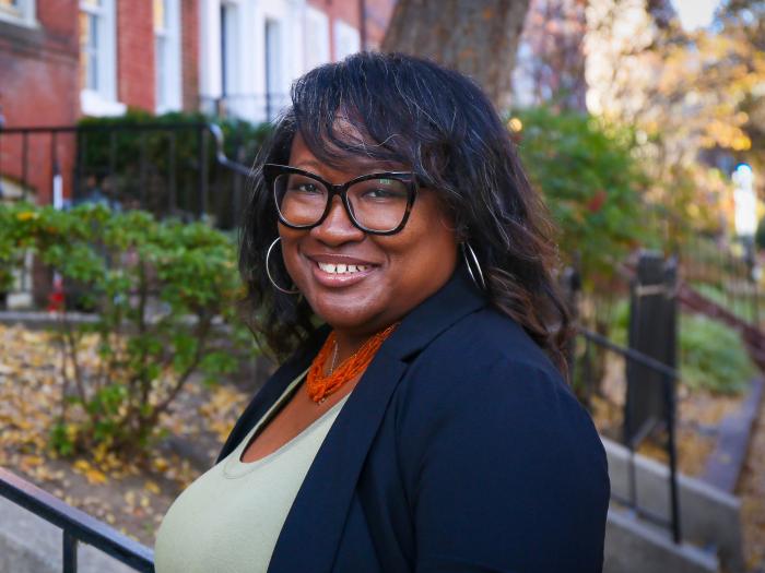 Headshot of Jameta Barlow in a green shirt with a blue jacket over it standing in front of a street of row houses.